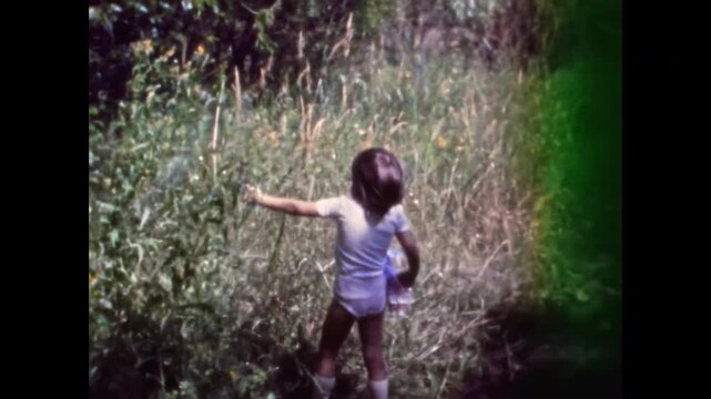 Archival film, charming little girl looks at flowers, playing in green, sunny nature of summer. Carefree and happy childhood memories. Sweet kid. Old film. Archival retro 1960s. Vintage archive.