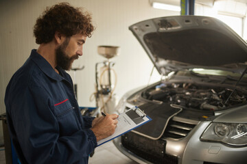 A mechanic wears a blue uniform and carefully checks the engine of a vehicle. He writes notes on a clipboard in a well-lit repair environment.