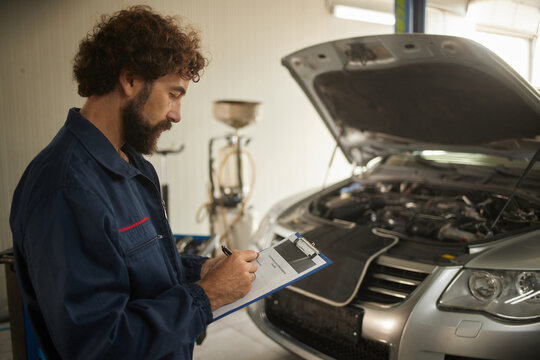 A mechanic wears a blue uniform and carefully checks the engine of a vehicle. He writes notes on a clipboard in a well-lit repair environment.