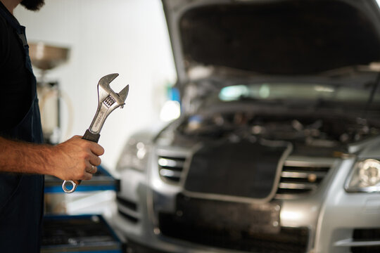 A mechanic holds a wrench while inspecting a car's engine in an auto shop. The vehicle's hood is open, revealing the engine components clearly.