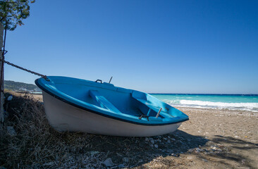 Blue fishing boat chained on sandy beach with turquoise sea waves and clear blue summer sky, Mediterranean coastal landscape, travel and vacation concept.
