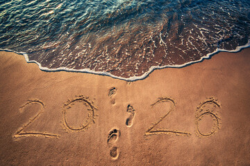 Happy New Year 2026 concept text written on the sea beach at sunrise and foot prints go to the sea water