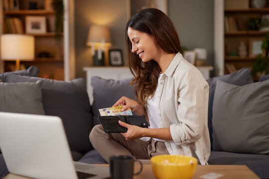 A woman is smiling as she sorts through cash in her wallet while seated comfortably on a couch. A laptop is open in front of her in a warm and inviting living room.