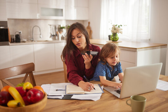 A mother and her child are engaged in activities at a kitchen table. The mother speaks on a phone while working on a notebook, and the child types on a laptop nearby.