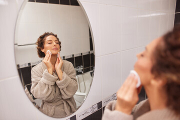 A woman in a cozy robe performs her daily skincare routine in a well-lit bathroom. She gently wipes...