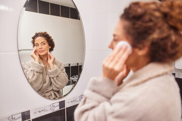 In a modern bathroom, a woman in a soft robe gently uses a cotton pad to cleanse her face while looking into the mirror, enjoying a relaxing moment of self-care. © Stockphotodirectors