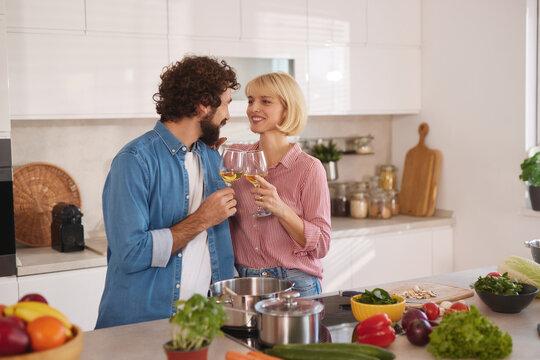 A couple stands close together in a bright kitchen, smiling and holding glasses of drink while preparing a meal with fresh vegetables and other ingredients on the counter. - Powered by Adobe