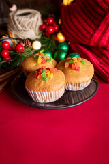 A warm and festive still life of Christmas-themed cupcakes topped with colorful candied fruit. The cupcakes are arranged with holiday decorations including pine branches, red berries, and rustic.