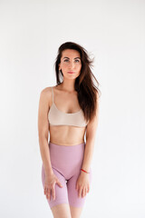 Fototapeta premium Sports woman standing in activewear with abs muscles, looking at camera isolated on white background. Caucasian 30s fitness girl wears beige top and pink leggings. Slim fit brunette with blue eyes.
