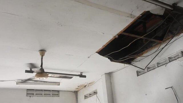 Rotating ceiling fan below severely damaged plasterboard with a structural hole exposing wiring and beams. Urgent home repair, leak, or renovation needed due to water damage 
