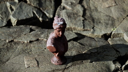 A small brown sculpture of a woman stands on a rough, rocky surface. Sunlight highlights the details while shadows create a striking contrast. This scene captures a moment of quiet beauty outdoors.