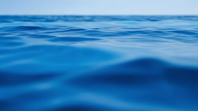 A serene, low-angle shot captures the tranquil surface of deep blue water, gently rippling under a soft, diffused sky. The calm, undulating waves create a soothing visual texture, suggesting peace, pu
