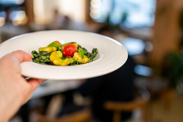 A hand holds a white plate featuring vibrant green spinach pasta with sautéed shrimp, fresh basil leaves, and a single cherry tomato, presented in a blurred restaurant setting