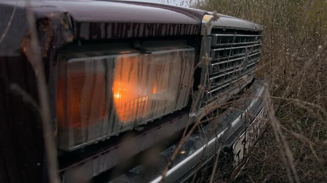 A close-up of the headlight of a Russian Zhiguli. The headlight comes on. The car is parked in a field among yellowed autumn grass