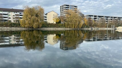 Residential buildings reflected on a still canal with autumn trees and overcast sky creating a...