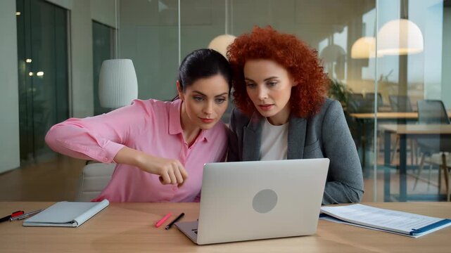 Two professional businesswomen are focusing on a laptop screen, discussing work and collaborating on a project during a productive meeting in a contemporary office environment