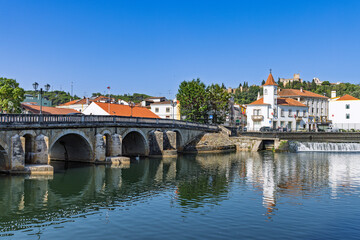 A scenic view of the River Nabao in Tomar, Portugal, featuring a historic arched stone bridge and the city's cityscape under a clear blue sky.