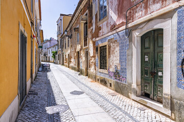 A charming, sun-dappled cobblestone alley in the historic center of Tomar, Portugal, featuring old buildings with faded tiles and a green wooden door.