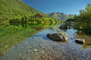 View of a serene lake reflecting the lush green mountains and a wooden chalet under a clear blue sky, Zelene pleso, Presovsky kraj, Slovakia.