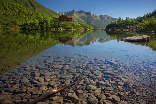 View of a tranquil lake mirroring a wooden cabin nestled amid green trees and distant mountains under a clear sky, Tatranska Javorina, Zelene pleso tarn, Presovsky kraj, Slovakia.