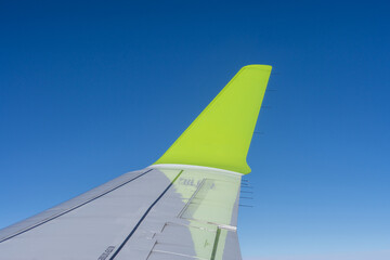 Airplane wing with bright green winglet against clear blue sky, view from passenger window during flight, concept of travel, tourism and modern aviation.