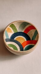 A close up overhead shot of a small, round ceramic bowl featuring a vibrant, abstract rainbow arc pattern with bold colors and a speckled off white glaze.