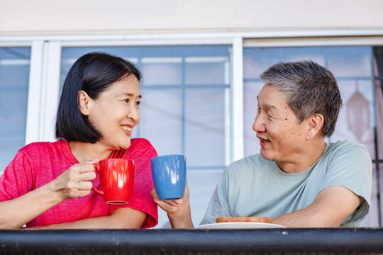 A middle-aged asian couple smiling together and cheerfully holding coffee mugs while having breakfast on their balcony at home.