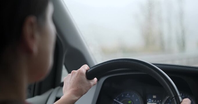 Over the shoulder view of a woman driving a car during heavy rain. Hands on steering wheel, windshield wipers clearing water from the glass. Bad weather driving.