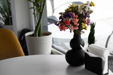 Close-up still life arrangement featuring a bouquet of colorful dried flowers in a modern black vase and napkin holder on a white table. Modern minimalist interior design - decor of cafe or restaurant