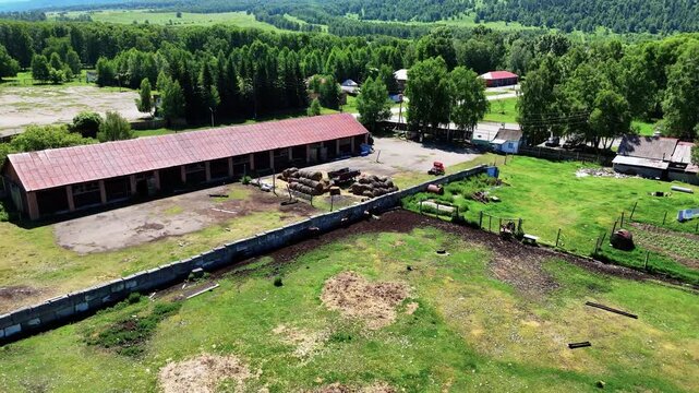 Drone pulls back from a building and an enclosure with grazing marals on a green meadow, with trees in the distance. Natural landscape of East Kazakhstan