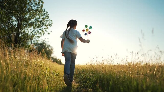 Child walks through grassy meadow at sunset holding colorful pinwheel toy while sunlight backlights braid and tall grass sways in field creating warm playful outdoor mood of summer light and motion