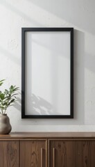 A blank black-framed picture frame on a wooden cabinet with a potted plant beside it. The wall is light-colored, creating a minimalist decor.