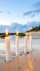 Four candles with lit flames on a sandy beach against a cloudy sky at sunset