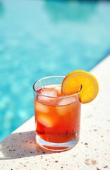 Obraz premium Glass of Negroni cocktail with orange slice on edge, served by swimming pool. Cocktail drink on stone surface with ice. Orange colored alcoholic beverage on vacation at luxury resort. Summer holiday,