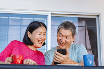 A happy middle-aged Asian couple watches on their phones and laughs together while eating breakfast toast and drinking coffee on a morning balcony.