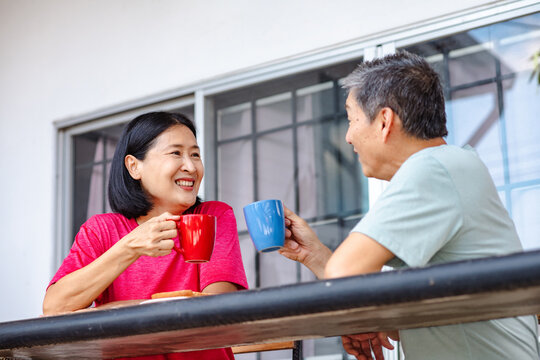 A happy middle-aged Asian couple enjoying breakfast on their balcony in the morning, smiling at each other and raising coffee mugs in a gesture of affection, highlighting a loving lifestyle. - Powered by Adobe
