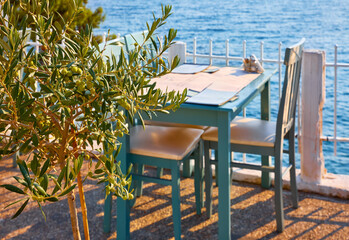 Sithonia, Chalkidiki, Greece. Traditional tavern cafe exterior with wooden table chairs. Olive tree branch focused in front with the view of the aegean sea in the background..