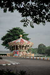 Classic colorful carousel merry-go-round in green park with tree framing and paved foreground.