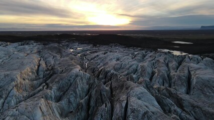 Aerial view of a rugged, grey glacier, contrasting with the dark, flat landscape under a bright, cloudy sky, creating a striking display of natural beauty, Iceland.