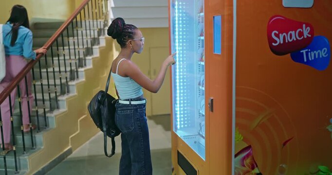 African happy young adult gen z female use easy fast self service choose juice drink press button campus area. male man walk stairway thirsty girl take bottle enjoy break day time hall way side view