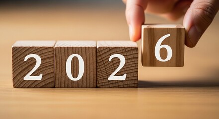 Changing the year 2026, marking a new beginning. A person's hand is seen adjusting a wooden block labeled '6'. This symbolizes change and the coming year, on a wooden surface