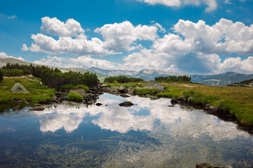 Scenic view of a high-altitude glacial lake in a mountainous region, with clouds reflected in the water. The landscape is typical of the Rila Mountain in Bulgaria. 