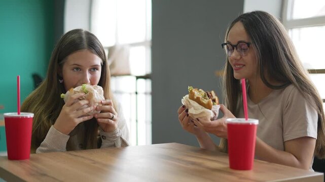 Two teenage girls eating tasty burger together at casual cafe restaurant table, holding wrapped burger and red drink cup with straw, sharing lunch and smiling while friend enjoys fastfood meal.