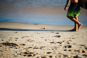 A surfer is seen walking barefoot on the edge of Barra beach during strong sunshine. Salvador, Bahia