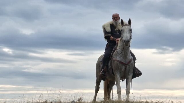 Aerial drone establishing or tracking shot of Bearded warrior in fur cloak riding a white horse across open landscape at sunrise, mountains in the background. Historical, rugged, adventurous scene