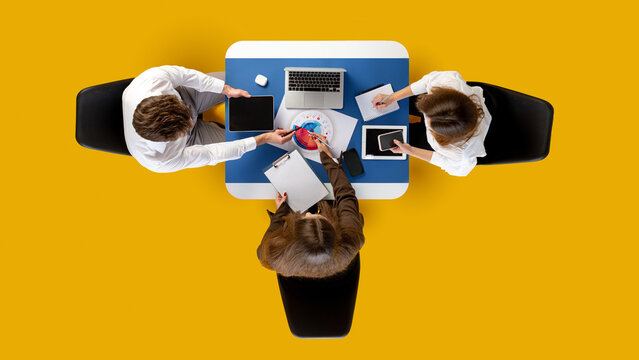 Team collaborating with digital devices around blue desk on yellow background in top-down view. Concept of corporate culture, business workflow, marketing communication and seminar materials.