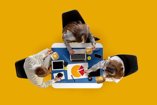 Team eating noodles while pointing to pie chart and using digital devices at desk. Concept of office culture, marketing workflow, food delivery and social media content.