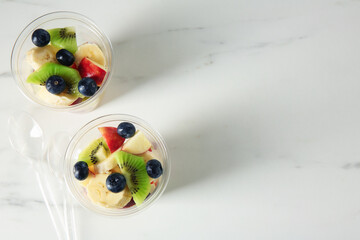 Tasty fruit salad in plastic cups and spoons on white marble table, flat lay. Space for text