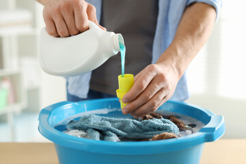 Man pouring detergent into cap over plastic basin with towels at table indoors, closeup
