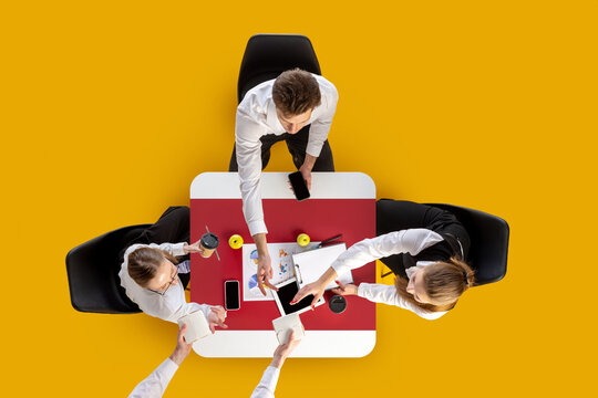 Office group discussing charts and exchanging take-away food boxes at red table. Concept of food delivery, corporate lifestyle, business workflow and startup collaboration.